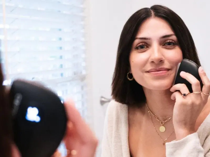 Gua sha tool used by smiling woman in front of mirror for facial toning and skincare routine.