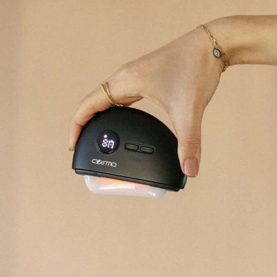 At home gua sha device held in hand with digital display and red light therapy on a neutral background.