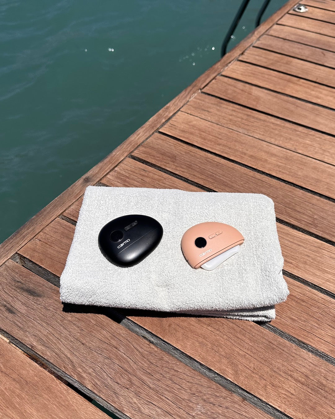Gua sha devices in black and peach resting on a towel beside a wooden dock overlooking calm water.