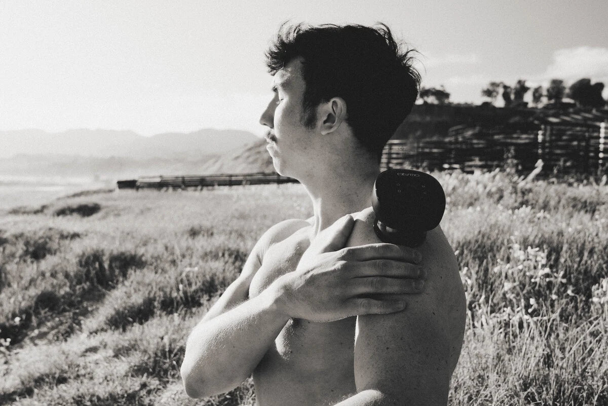 Smart cupping device applied to man's shoulder as he stands shirtless in a grassy outdoor field with distant mountains.