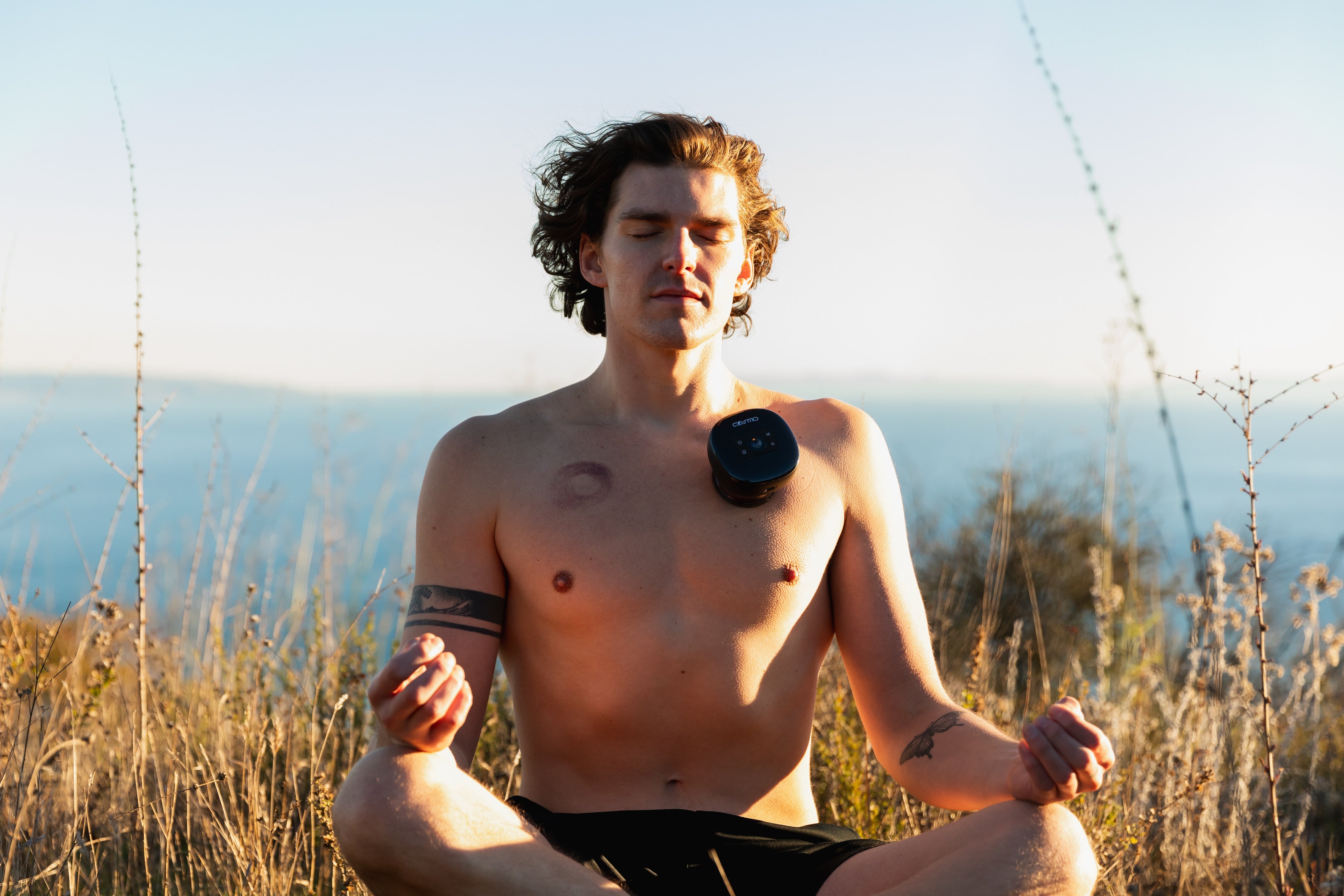 Cupping therapy machine applied to man’s chest as he meditates shirtless outdoors with ocean view in the background.