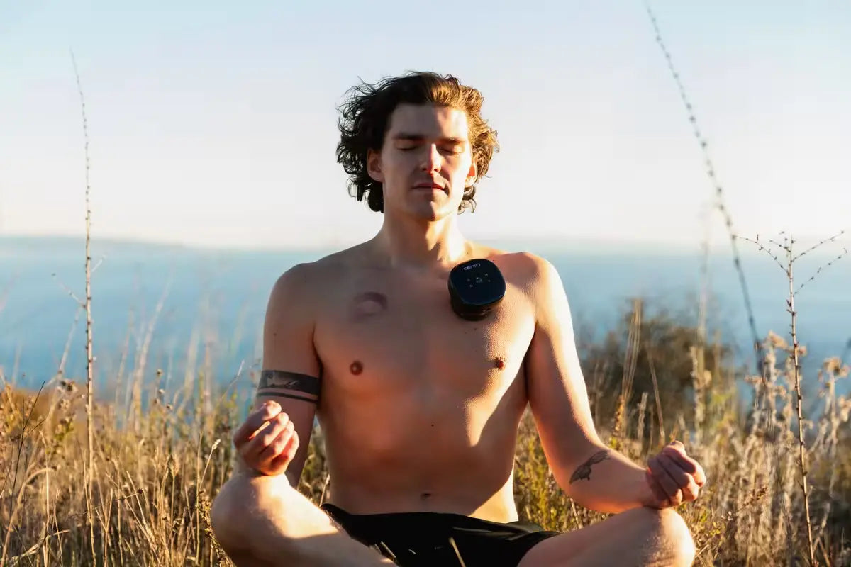 Cupping therapy machine applied to man’s chest as he meditates shirtless outdoors with ocean view in the background.