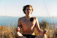 Cupping therapy machine applied to man’s chest as he meditates shirtless outdoors with ocean view in the background.