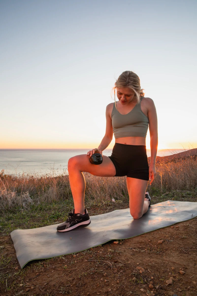 Smart cupping therapy device used on thigh by woman kneeling on yoga mat during sunset near the ocean.